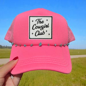 May include: A bright pink trucker hat with a white square patch that reads "The Cowgirl Club" in black script. The hat has a mesh back and a curved brim. Turquoise-colored stones are attached to the brim.