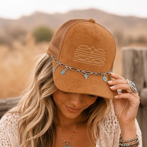 May include: A tan suede trucker hat featuring a mesh back and a white embroidered geometric design on the front panel. A silver-toned metal chain is attached across the brim, adorned with small turquoise-colored charms shaped like flowers.