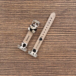 May include: Two watch straps with a cow print design. One strap is engraved with the name "Emma", and the other has the date "12.12.1998" and a heart symbol. The straps are made of a light-colored material with black and white cow print accents.