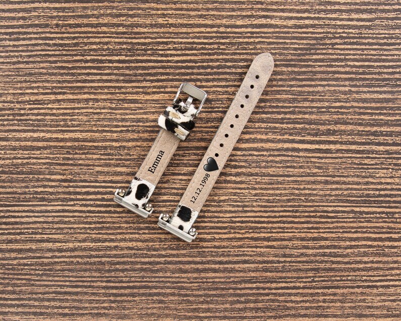 May include: Two watch straps with a cow print design. One strap is engraved with the name "Emma", and the other has the date "12.12.1998" and a heart symbol. The straps are made of a light-colored material with black and white cow print accents.
