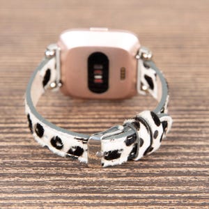 May include: A rose gold smartwatch with a black screen and a white and black spotted leather band. The band is secured with a silver buckle. The watch is displayed on a wooden surface.