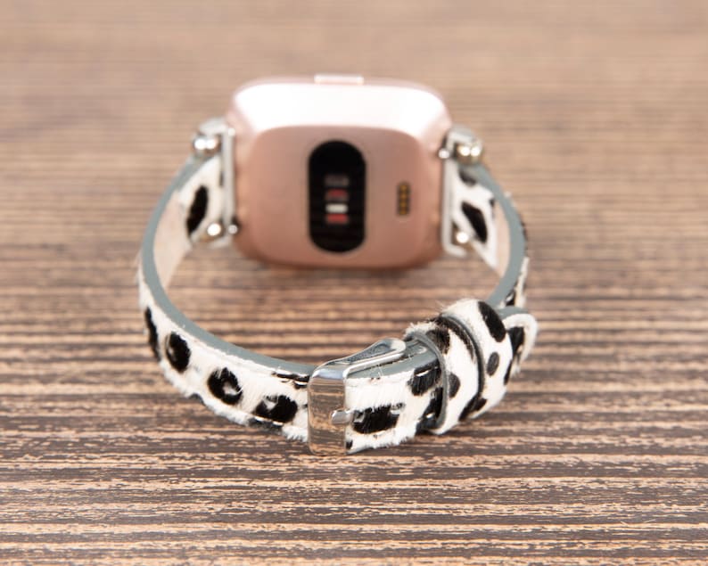 May include: A rose gold smartwatch with a black screen and a white and black spotted leather band. The band is secured with a silver buckle. The watch is displayed on a wooden surface.