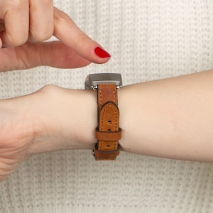 Könnte beinhalten: Eine quadratische Smartwatch mit braunem Lederarmband wird am Handgelenk getragen. Das Armband wird mit einer Schnalle befestigt. Das Zifferblatt der Uhr hat eine dunkle Metallfarbe. Ein Finger mit rotem Nagellack zeigt auf die Uhr.