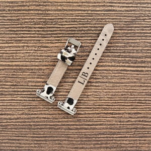 May include: Two-piece watch strap set. The straps are light beige with black and white cow print accents. The buckle is silver. The strap has the initials "L/B" engraved on it.