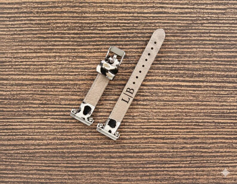 May include: Two-piece watch strap set. The straps are light beige with black and white cow print accents. The buckle is silver. The strap has the initials "L/B" engraved on it.