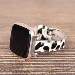 May include: A rose gold smartwatch with a black screen and a patterned watch band. The band features a black and white spotted design. The watch is resting on a wooden surface.