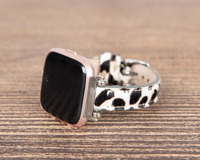 May include: A rose gold smartwatch with a black screen and a patterned watch band. The band features a black and white spotted design. The watch is resting on a wooden surface.