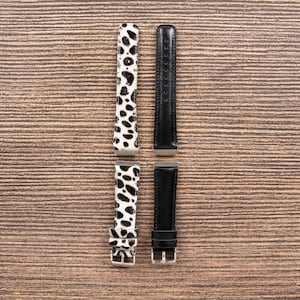 May include: Four watch straps are displayed against a wood grain background. Two straps feature a black and white spotted pattern, while the other two are solid black. The straps have silver-colored buckles and connectors.