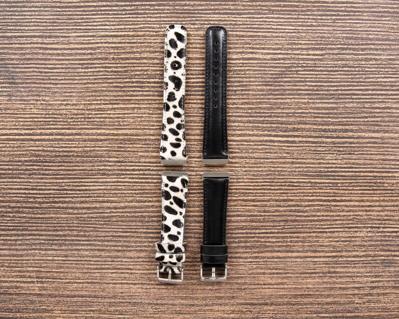 May include: Four watch straps are displayed against a wood grain background. Two straps feature a black and white spotted pattern, while the other two are solid black. The straps have silver-colored buckles and connectors.