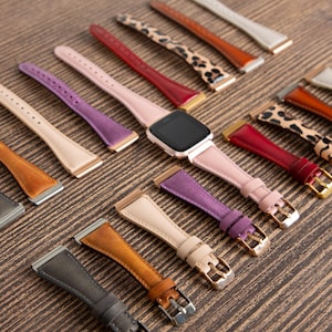 May include: A collection of watch bands in various colors and patterns, including black, brown, purple, and leopard print. A smartwatch with a black screen is in the center, surrounded by the bands. The bands have metal buckles and are arranged on a wooden surface.