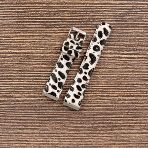 May include: Two watch straps with a black and white spotted pattern, resembling a Dalmatian's coat. The straps have silver-colored buckles and are laid out on a brown wooden surface.