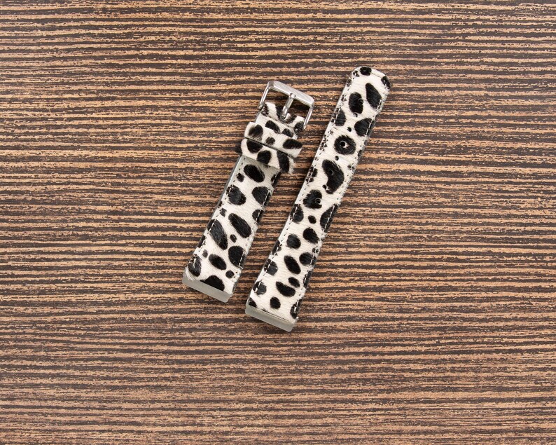 May include: Two watch straps with a black and white spotted pattern, resembling a Dalmatian's coat. The straps have silver-colored buckles and are laid out on a brown wooden surface.