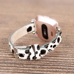 May include: A smartwatch with a rose gold case and a black and white spotted leather band. The band has a silver buckle and is attached to the watch with silver pins. The watch is resting on a wooden surface.