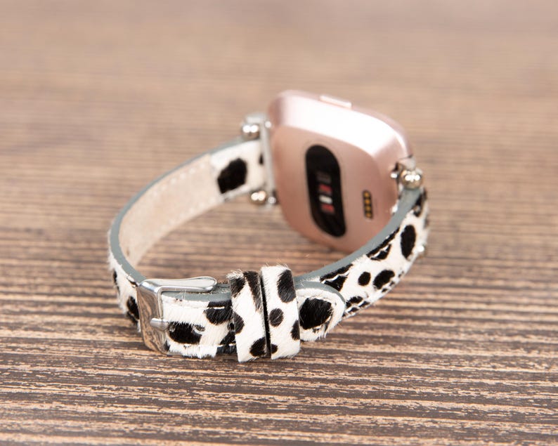 May include: A smartwatch with a rose gold case and a black and white spotted leather band. The band has a silver buckle and is attached to the watch with silver pins. The watch is resting on a wooden surface.