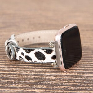 May include: A rose gold smartwatch with a black screen and a black and white cowhide patterned watch band. The band is made of leather and has a light beige underside. The watch is on a wooden surface.