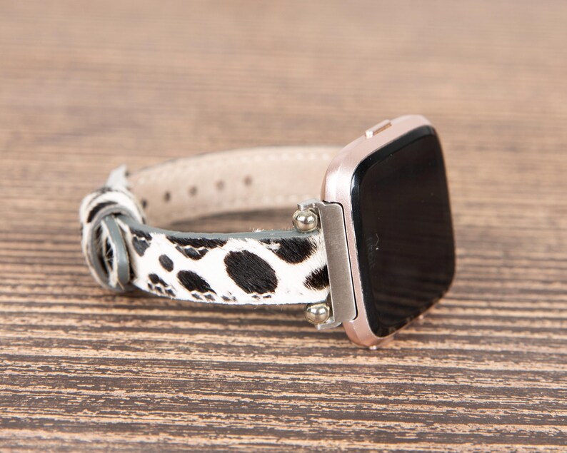 May include: A rose gold smartwatch with a black screen and a black and white cowhide patterned watch band. The band is made of leather and has a light beige underside. The watch is on a wooden surface.