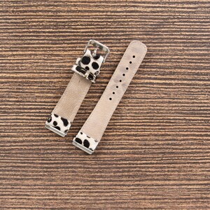 May include: Two-piece watch strap with a light brown leather band and black and white spotted accents. The strap has a silver-colored buckle and several holes for adjustment. The watch strap is displayed on a wooden surface.