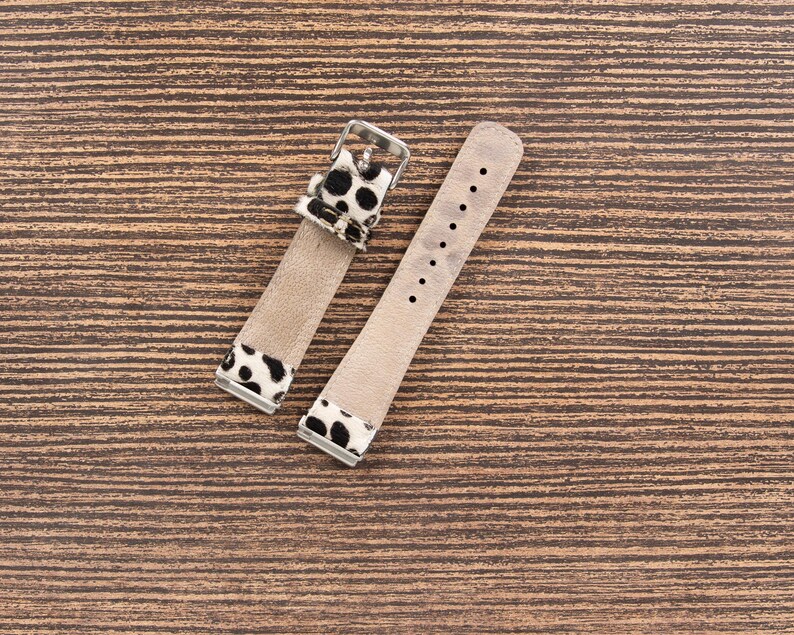 May include: Two-piece watch strap with a light brown leather band and black and white spotted accents. The strap has a silver-colored buckle and several holes for adjustment. The watch strap is displayed on a wooden surface.