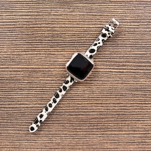 May include: A smartwatch with a black screen and a white and black spotted band. The watch case is silver. The band is made of a leather-like material and is secured with a silver buckle. The watch is lying on a brown wooden surface.