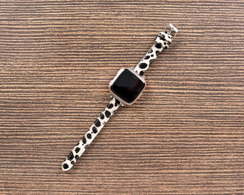May include: A smartwatch with a black screen and a white and black spotted band. The watch case is silver. The band is made of a leather-like material and is secured with a silver buckle. The watch is lying on a brown wooden surface.