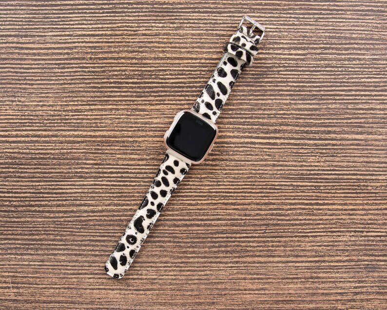 May include: A smartwatch with a black screen and a white and black spotted band. The watch has a silver-colored frame and a silver buckle. The band is made of a material with a pattern of black spots on a white background.