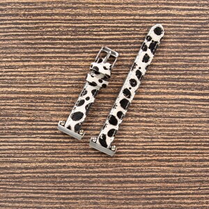 May include: Two-piece watch band with a black and white spotted pattern. The band has silver-colored metal hardware, including a buckle and connectors. The watch band is made of a textured material and is set against a brown wooden background.