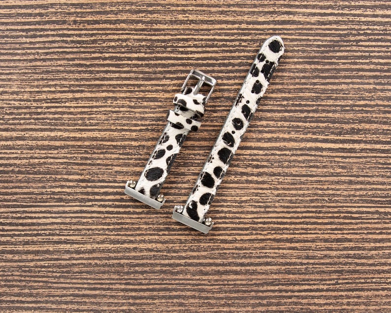 May include: Two-piece watch band with a black and white spotted pattern. The band has silver-colored metal hardware, including a buckle and connectors. The watch band is made of a textured material and is set against a brown wooden background.