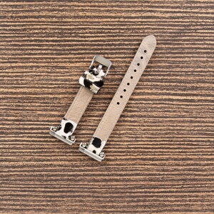 May include: Two-piece watch band with a light beige leather strap and silver-tone metal hardware. The band features a black and white cow print accent near the buckle and attachment points. The band is designed to fit a watch.