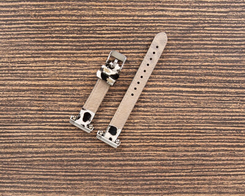 May include: Two-piece watch band with a light beige leather strap and silver-tone metal hardware. The band features a black and white cow print accent near the buckle and attachment points. The band is designed to fit a watch.