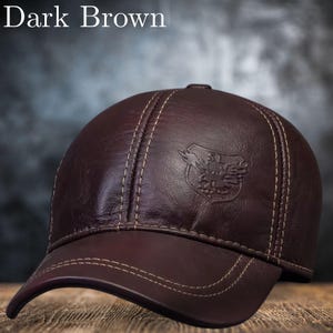 Könnte beinhalten: Dunkelbraune Baseballkappe aus Leder mit gebogenem Schirm und Ziernähten. Die Kappe hat ein dekoratives Emblem und den Text "Dark Brown".