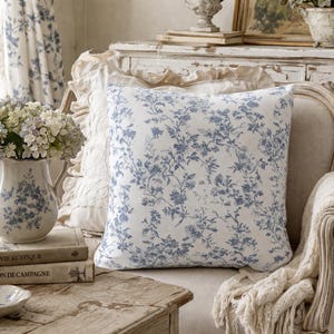 May include: A square cushion with a blue floral design on a white backdrop. The cushion is on a cream-coloured chair with a white fringed throw. A matching vase with flowers and books are on a wooden table.