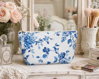 May include: A white cosmetic bag with a blue floral pattern, featuring roses and foliage. The bag has a gold zipper and is displayed on a white dresser. A vase of pink roses and makeup brushes are in the background.