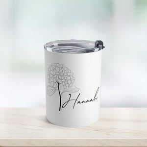 Op de afbeelding: Witte beker met een zwarte bloemenlijntekening en de naam "Hannah" in cursief schrift. De beker heeft een doorzichtig deksel met een zwarte klapsluiting. Het ontwerp is eenvoudig en elegant.