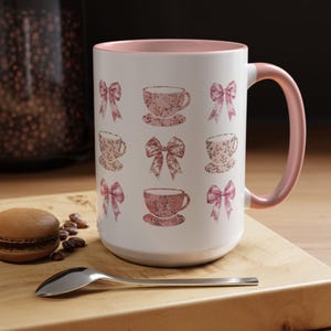 May include: A white ceramic mug with a pink rim and handle. The mug features a repeating pattern of pink teacups and bows with floral designs.