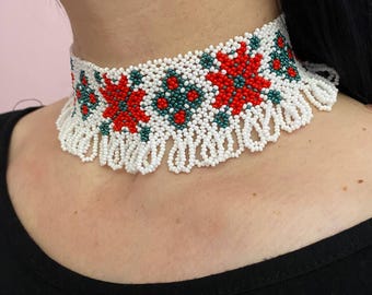 Może przedstawiać: Biała koralikowa choker z czerwonymi i zielonymi wzorami kwiatowymi. Naszyjnik ma frędzle z koralików na dole.