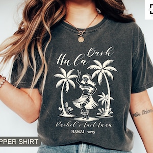May include: Dark gray t-shirt with a white graphic of a hula dancer, palm trees, and the text "Hula Bash". The text "Rachel's last luau" and "HAWAII - 2025" are also printed on the shirt. The shirt has a "Pepper Shirt" label.