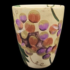 Könnte beinhalten: Cremefarbene Keramikvase mit handgemaltem Traubenmuster. Die Trauben sind in Lila, Orange und Gelb gehalten, mit grünen Blättern und Ranken. Die Vase hat eine leicht konische Form und eine glänzende Oberfläche.