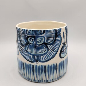 May include: A cylindrical ceramic planter with a cream-colored base and intricate blue floral patterns. The design features stylized flowers and vertical lines, creating a decorative and artistic look. The planter is suitable for indoor or outdoor use.