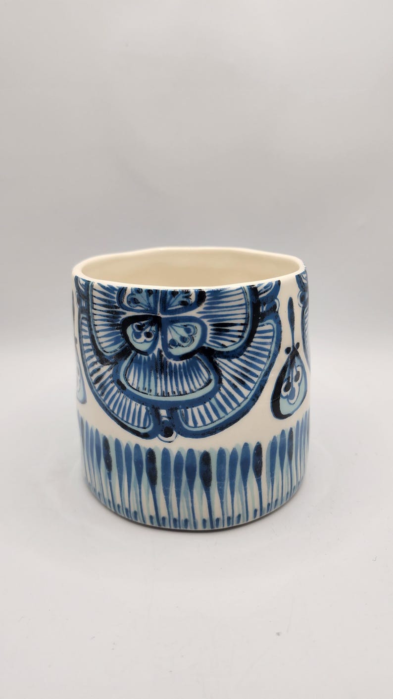 May include: A cylindrical ceramic planter with a cream-colored base and intricate blue floral patterns. The design features stylized flowers and vertical lines, creating a decorative and artistic look. The planter is suitable for indoor or outdoor use.