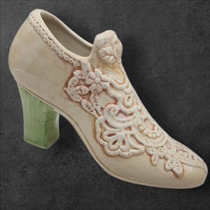 May include: Cream-colored ceramic shoe with a green heel. The shoe features intricate floral lace detailing and a decorative design on the vamp. The heel is a solid, light green color. The shoe is a decorative item.