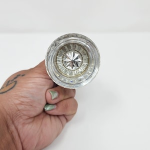 May include: A clear glass door knob featuring a circular shape with a faceted, star-like pattern at its center. The knob has a smooth, rounded outer edge and a reflective, metallic-toned base visible through the transparent glass material.
