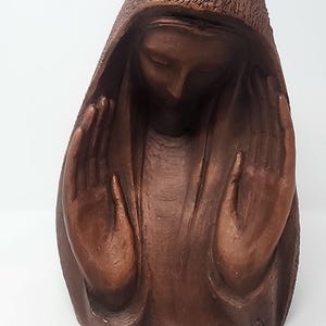 May include: A brown, monochromatic sculpture depicting a figure with a hooded head and downcast eyes. The figure has both hands raised with palms facing inward toward the face, featuring a textured surface finish throughout the entire piece.