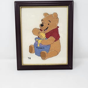 May include: A framed cross-stitch embroidery featuring a light brown bear wearing a red shirt, sitting and eating yellow honey from a blue pot labeled HUNNY. The initials TB are stitched in the lower left corner, all set in a dark brown frame.