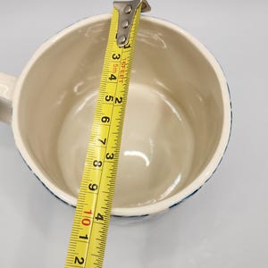 May include: A white ceramic mug with a blue patterned rim is measured with a yellow tape measure. The tape measure shows the mug is approximately 4 inches in diameter. The mug has a handle.