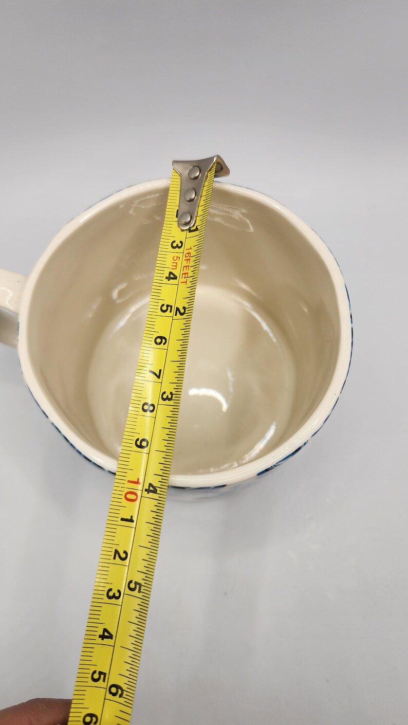 May include: A white ceramic mug with a blue patterned rim is measured with a yellow tape measure. The tape measure shows the mug is approximately 4 inches in diameter. The mug has a handle.