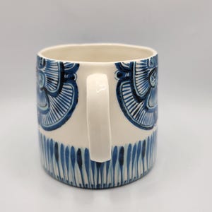 May include: A ceramic mug with a white handle and a cream-colored body. The mug is decorated with blue floral patterns and vertical blue lines near the base. The mug is designed for hot beverages.
