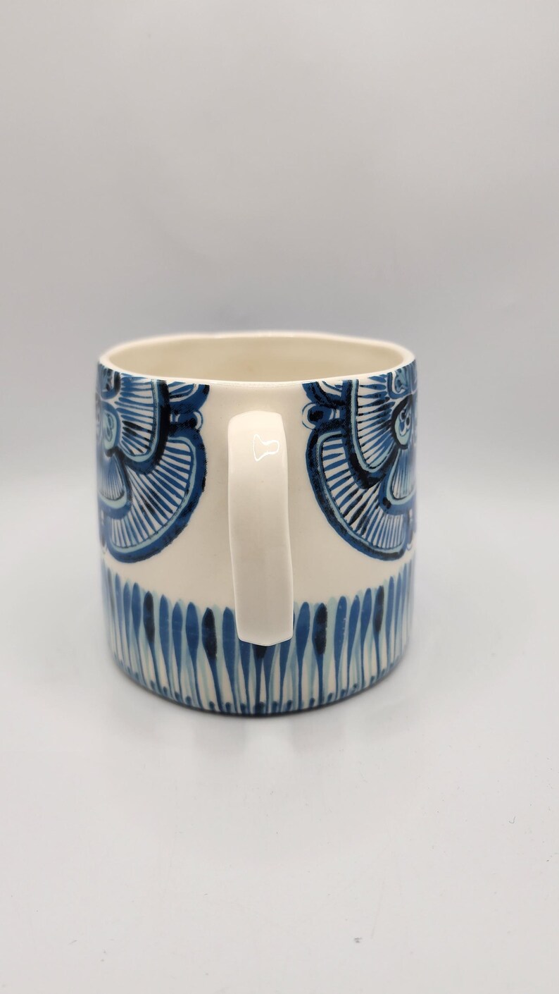 May include: A ceramic mug with a white handle and a cream-colored body. The mug is decorated with blue floral patterns and vertical blue lines near the base. The mug is designed for hot beverages.