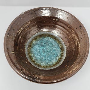 Peut inclure: Un bol en céramique fait à la main avec un extérieur marron et un intérieur turquoise. Le bol a une surface texturée et un large bord. Le centre du bol présente un design unique et mousseux. Ce bol décoratif est parfait pour servir ou exposer.