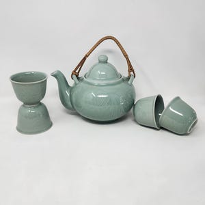 Peut inclure: Un service à thé en céramique vert céladon composé d'une théière avec un bec verseur incurvé, un couvercle et une anse tressée marron, accompagné de quatre tasses assorties. La théière et les tasses présentent des motifs de poissons en relief.