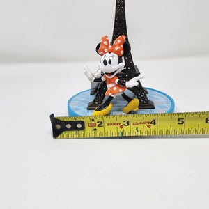 Pu&ograve; includere: Una statuetta in miniatura della Torre Eiffel con il personaggio di Minnie Mouse, che indossa un vestito arancione a pois bianchi e un fiocco abbinato. Un metro a nastro giallo mostra i centimetri.